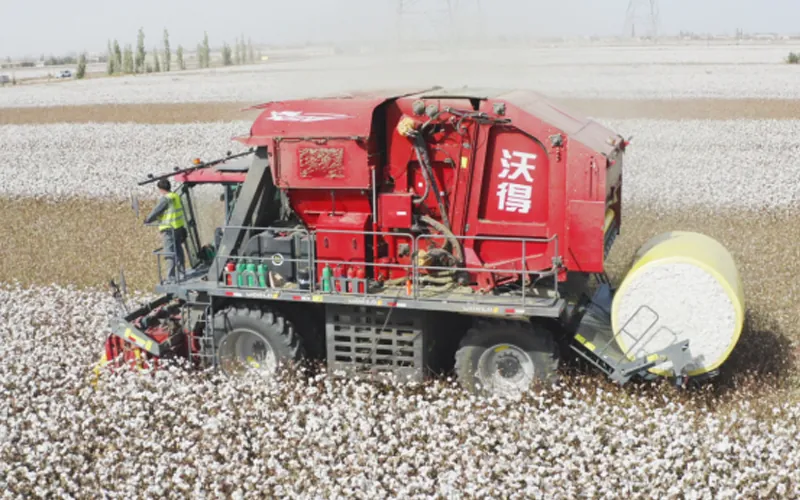 1 A red cotton harvester with the Chinese brand name "FMWorld" is working in a cotton field with a large round bale of harvested cotton attached.